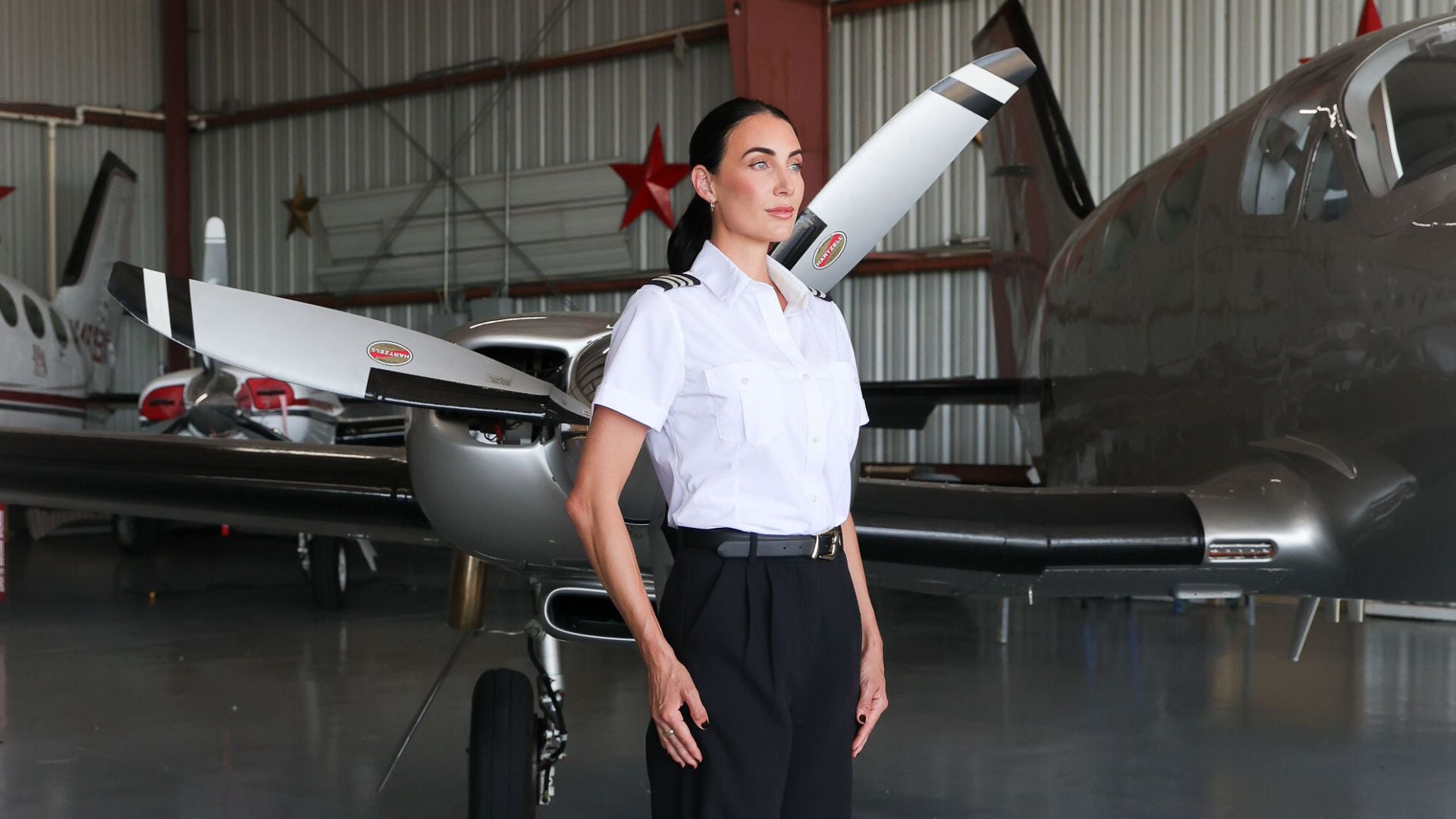 person standing in small airplane hangar near small aircraft, wearing  Skyshield's Women's Signature Short Sleeve pilot uniform shirt with black pants