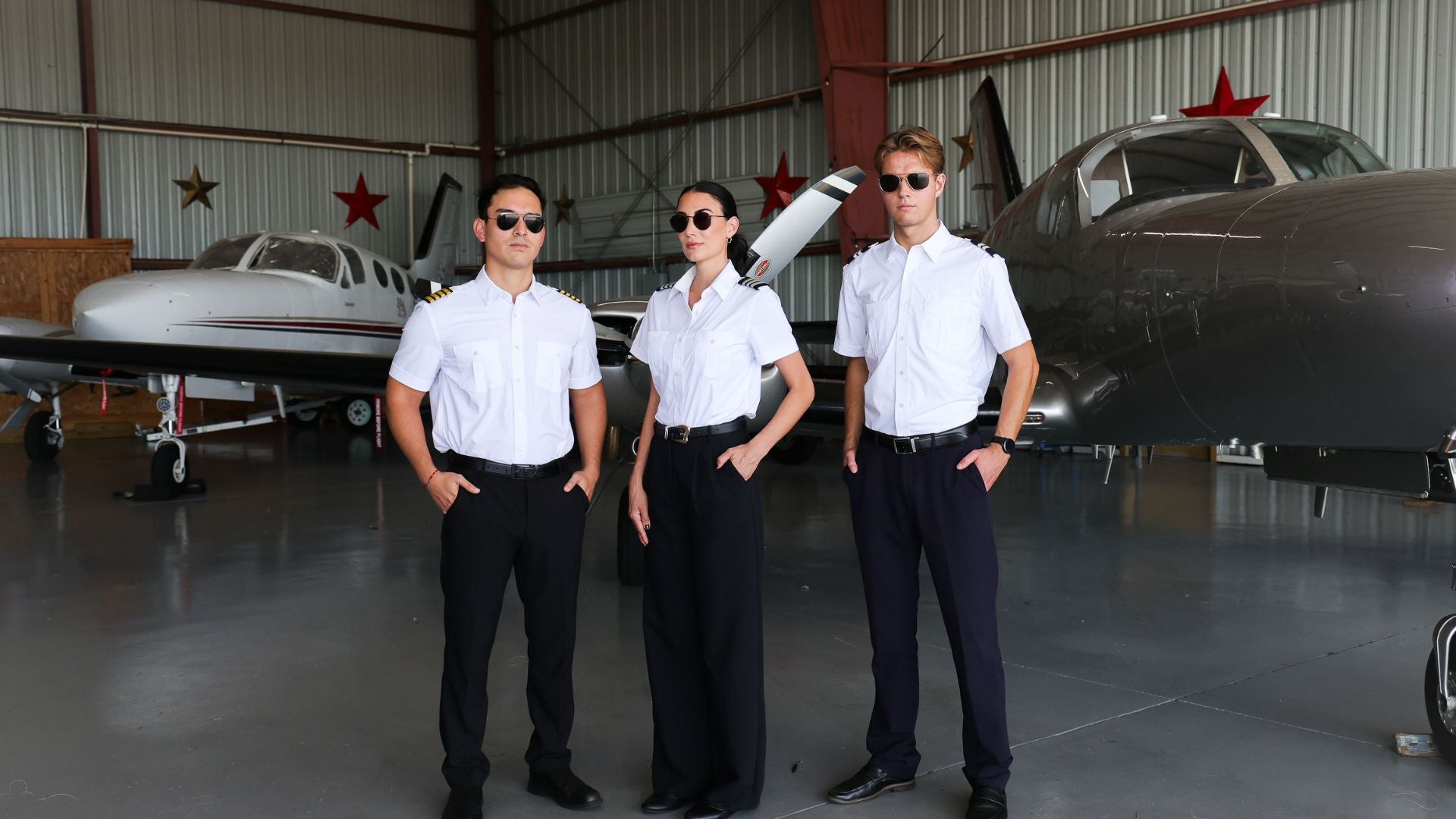 Three people wearing sunglasses and pilot uniforms, including white Women's and Men's Skyshield shirts with built-in UV protection, standing together inside an aircraft hangar with airplanes in background.